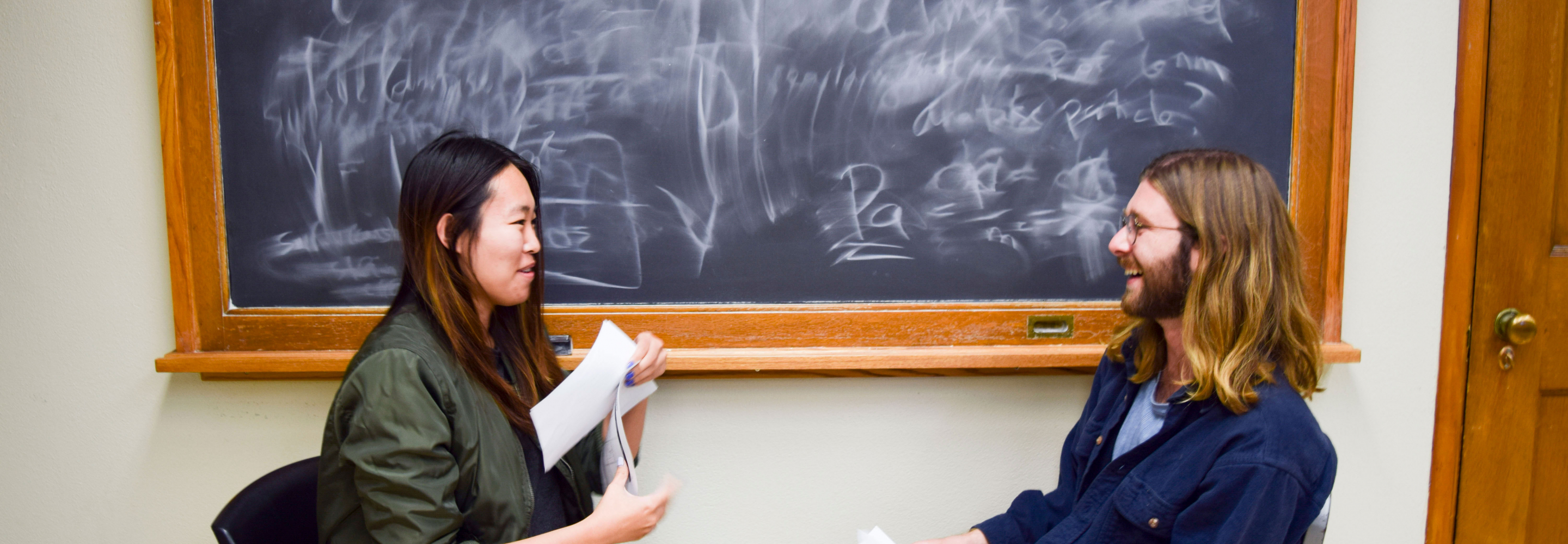 Two students sit in front of a blackboard talking