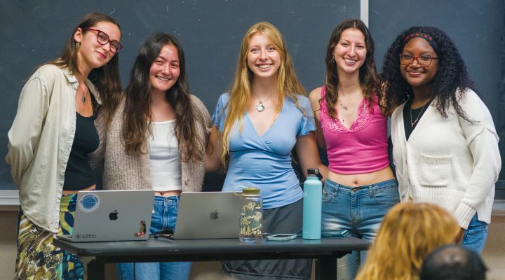 A group of young women in a classroom standing in a group in front of the chalkboard smiling