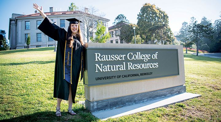 A woman in graduate cap and gown by a sign that says Rausser College of Natural Resources 