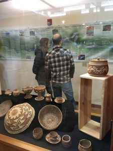 a man and woman with backs to camera examine a Karuk Tribe geographical history poster. Baskets in foreground.