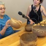 At right, a woman in a black sleevless top sits at microphone with pen and pad. At lef, a woman in a blue shirt sits at second mic holding a deep bowl-shaped woven tribal basket. Three other baskets in different sizes are on the table in front of them.