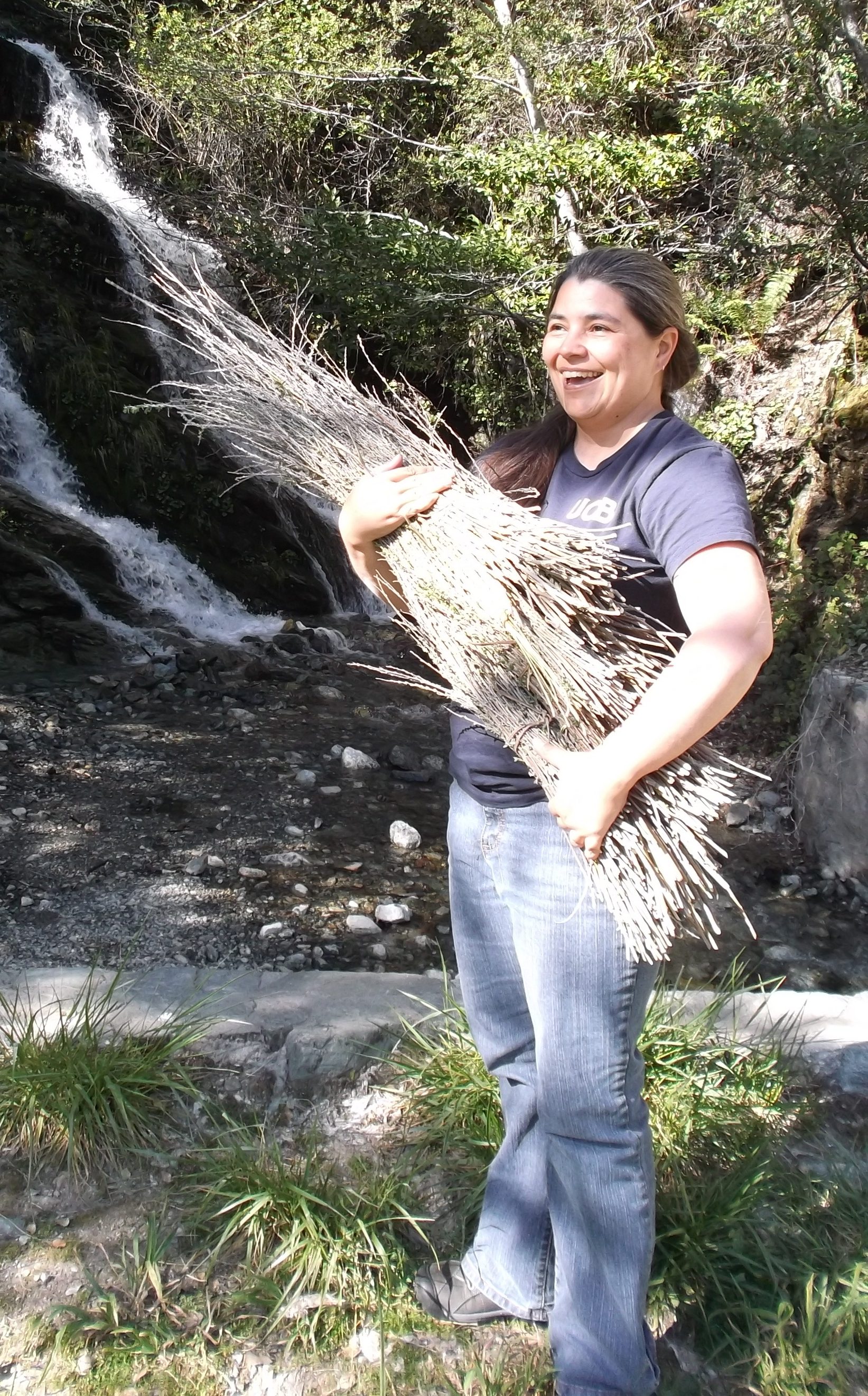 Carolyn Smith stands in front of a tree on a grassy area holding a sheaf of fiber used for basketry