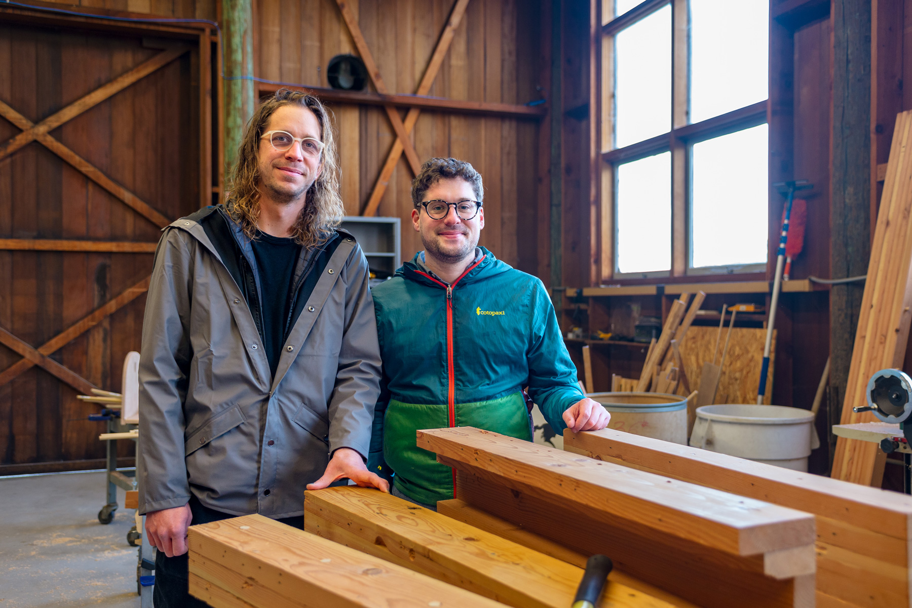 Two men stand in a wood shop and smile at the camera. A pile of lumber is on a table in front of them.
