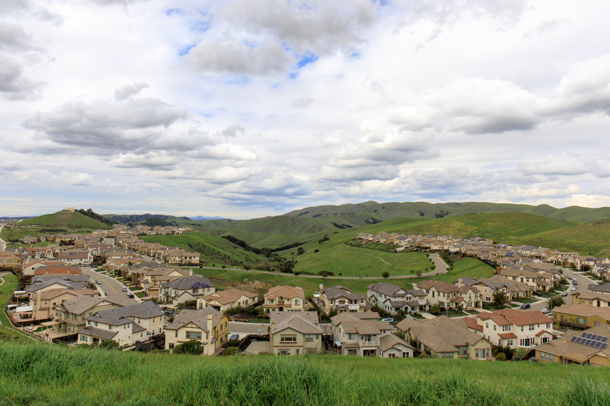 A suburban neighborhood with numerous houses is set against a backdrop of green hills and a cloudy sky.