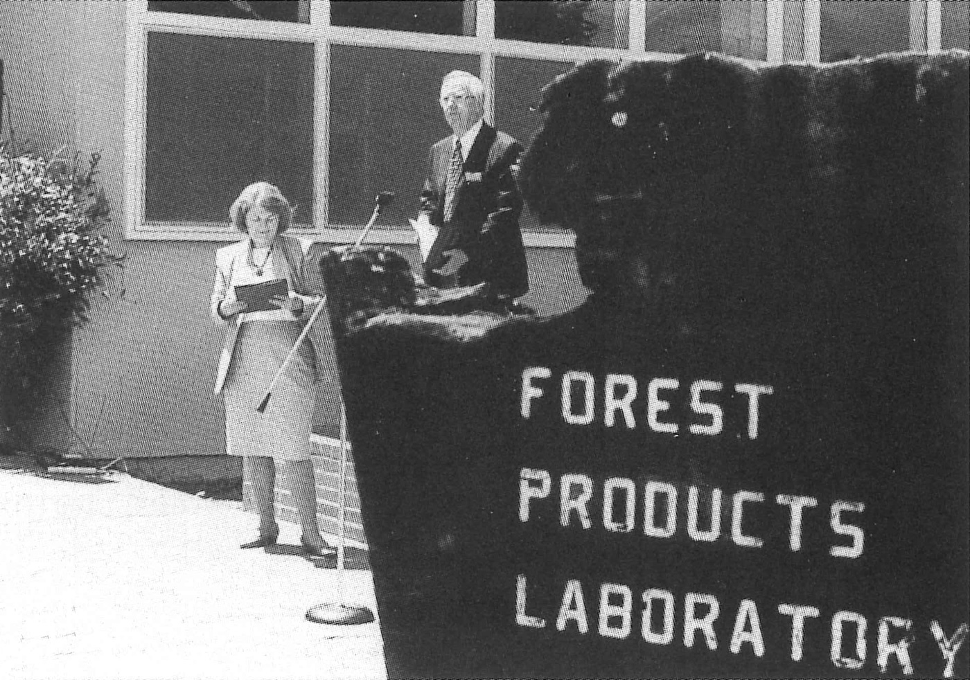 A black and white photo of a man standing at a lectern outdoors. A sign that reads "Forest Products Lab" is visible in the foreground.