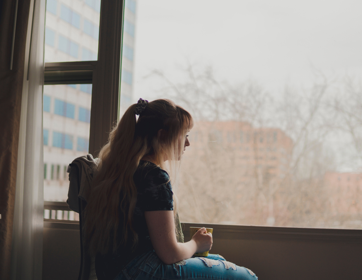 A young woman rests in a chair and looks out the window. The woman is holding a mug.