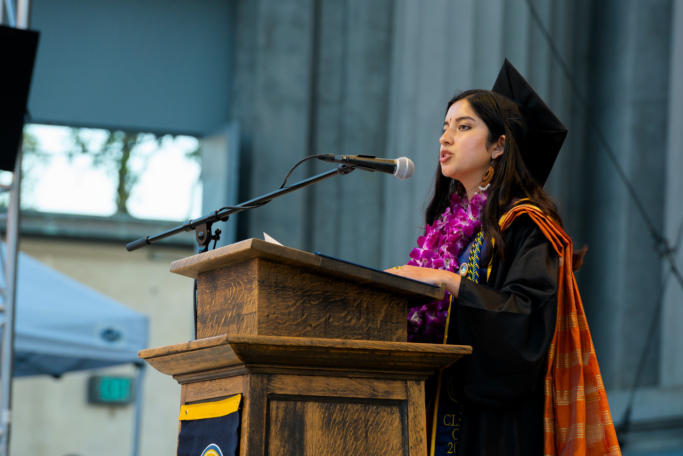 A photo of a woman in graduation regalia speaking while standing at a podium