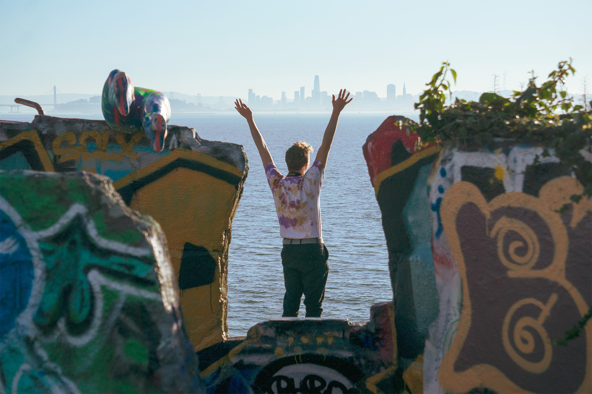 A person standing looking away from the camera with arms overhead. graffiti on concrete next to them and San Francisco skyline in the background.