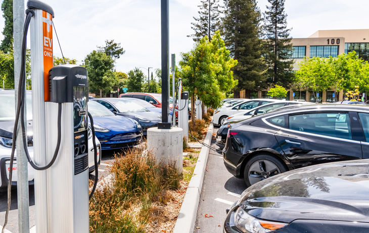 Electric cars charging in a parking lot with charging stations under a partly cloudy sky. Trees and a building are in the background.