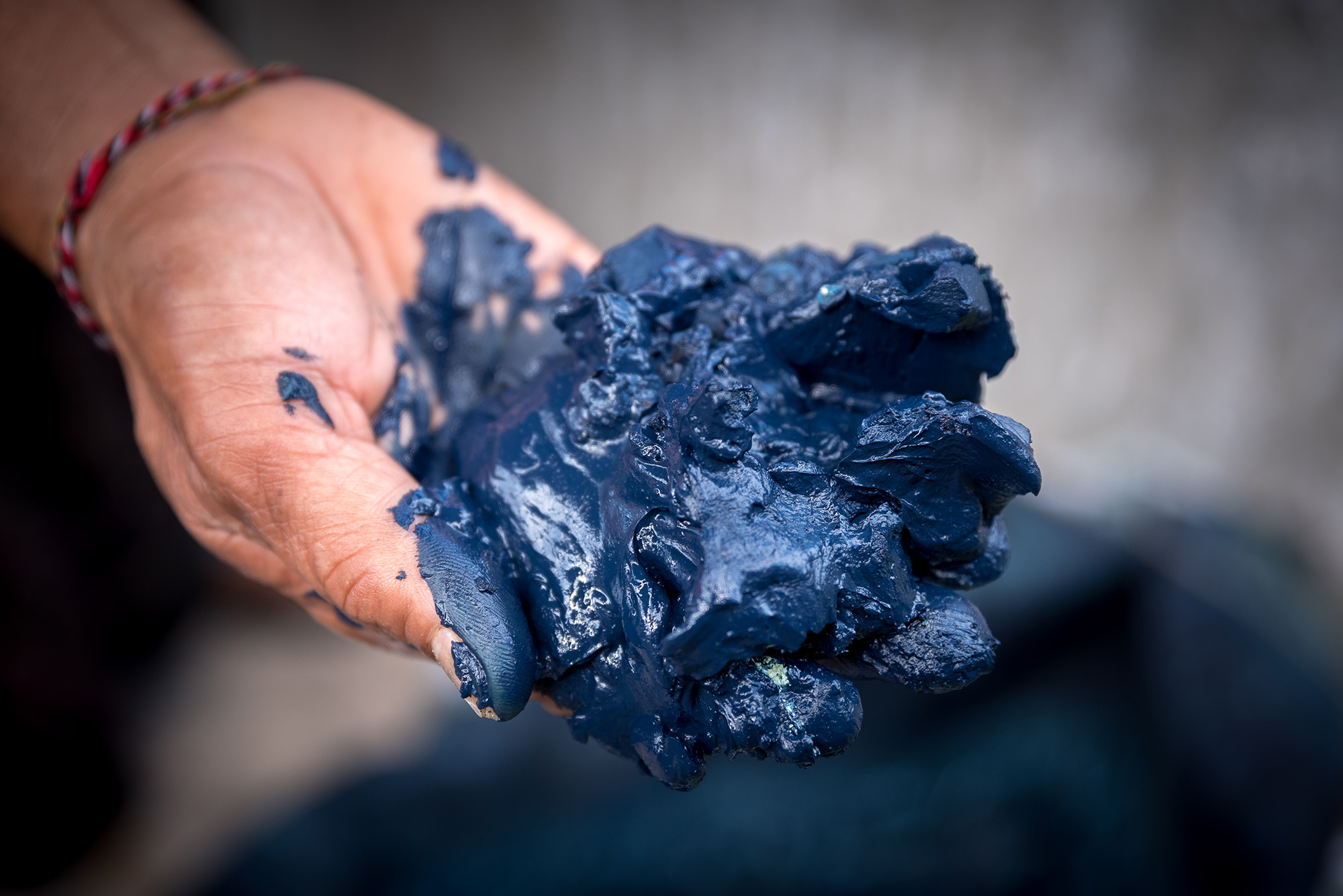 A hand with a string bracelet holds out indigo dye paste, which looks like wet, blue clay.