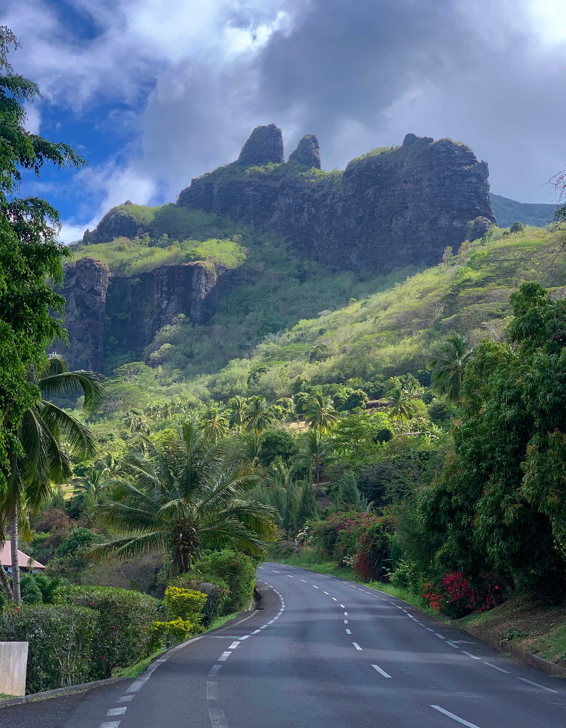 An empty paved road surrounded by a lush tropical mountainous terrain behind a clear sky.