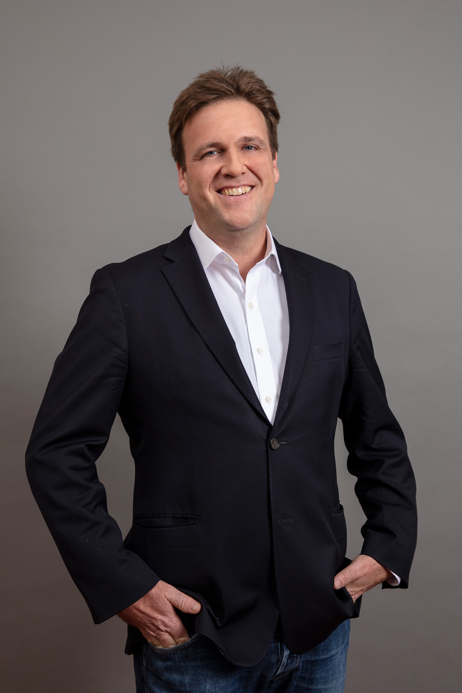 A headshot of a man standing against a grey background. The man is wearing a dark blazer and white shirt, and has his hands in his pockets.