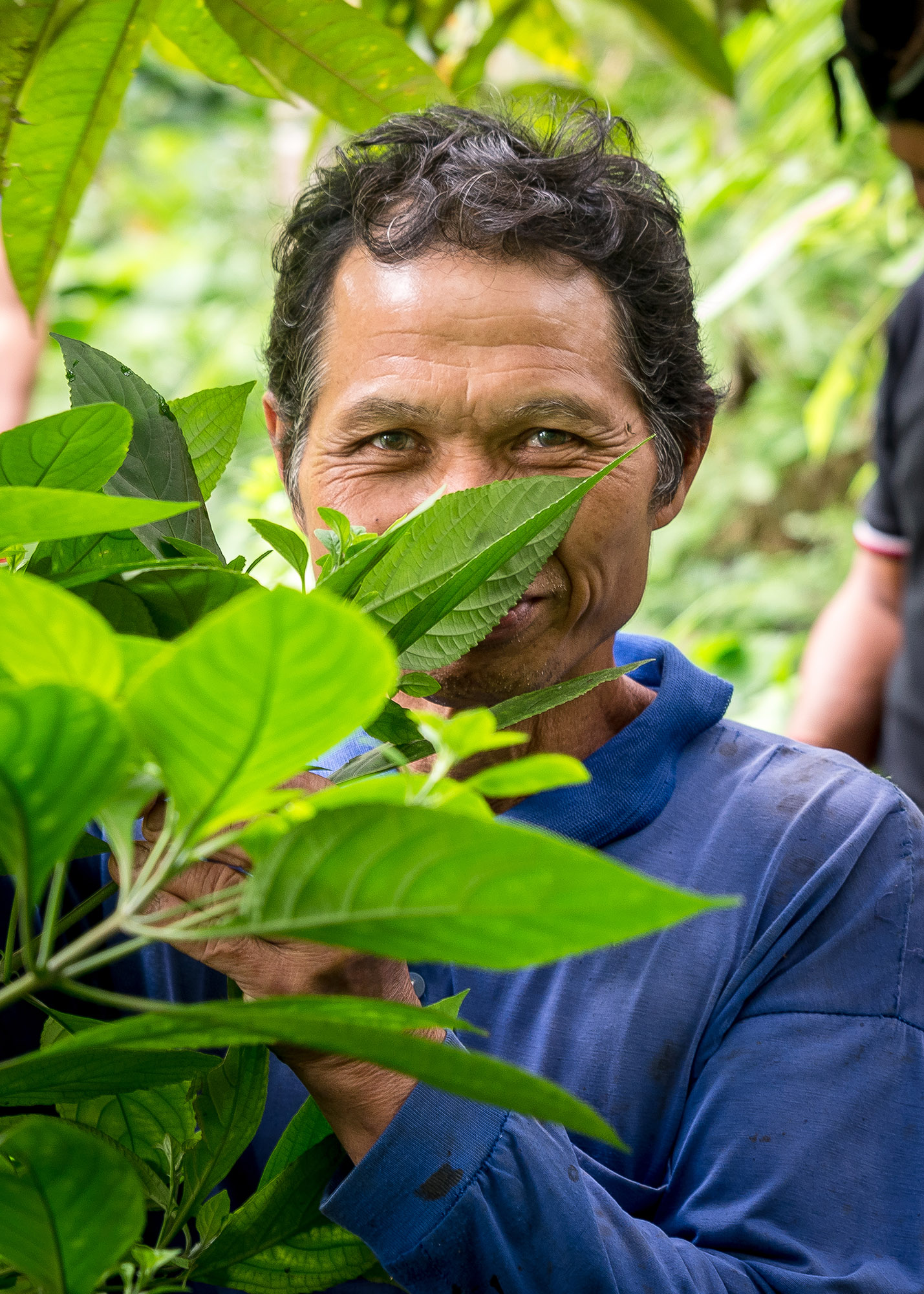 A man with tan skin and dark curly hair stands smiling behind leaves. His face is partially hidden, but you can see the smile-crinkles around his eyes.