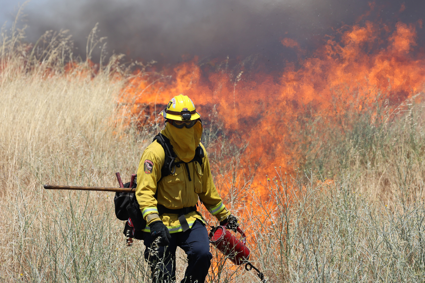 A person in yellow protective gear lights a fire in tall grass.