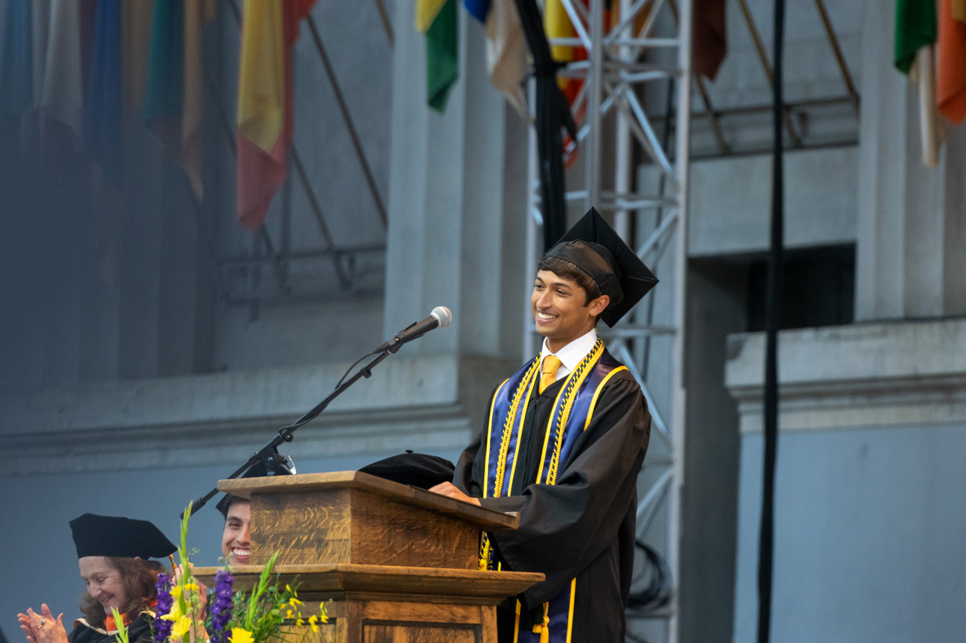 A photo of a man wearing graduation regalia smiling from behind a lectern.