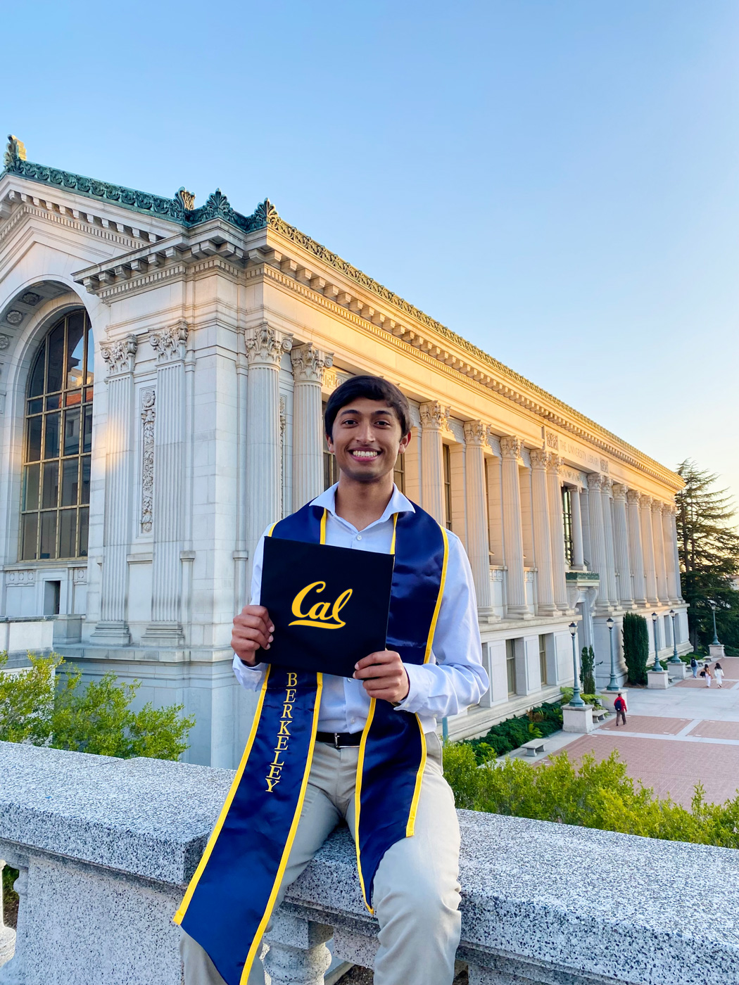 A photo of a young an sitting on a stone ledge in front of a building smiling. He holds a piece of paper that reads "Cal" written in script.
