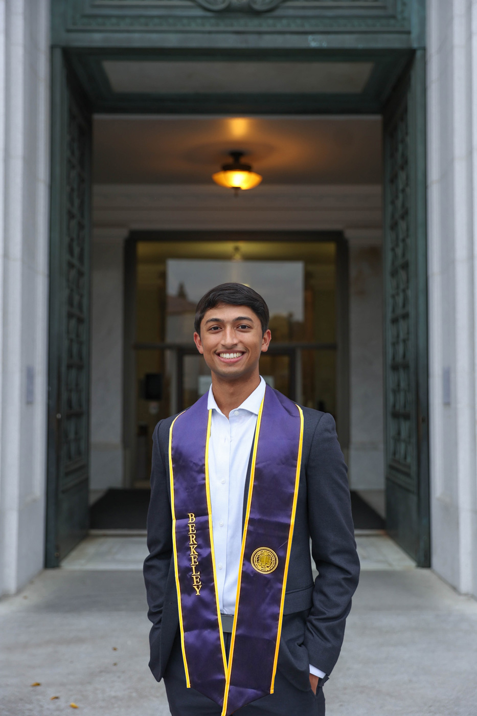 A photo of a smiling man wearing graduation regalia posing for a photo outside of a stone building.