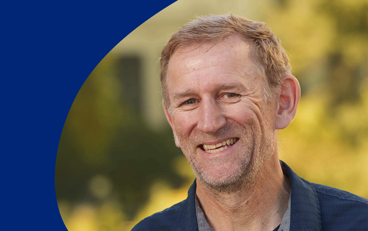 A graphic of a man in a dark blue collared shirt smiling at the camera. There is a medium blue swirl to his left, and the background image of trees and a building has a yellow color overlay.