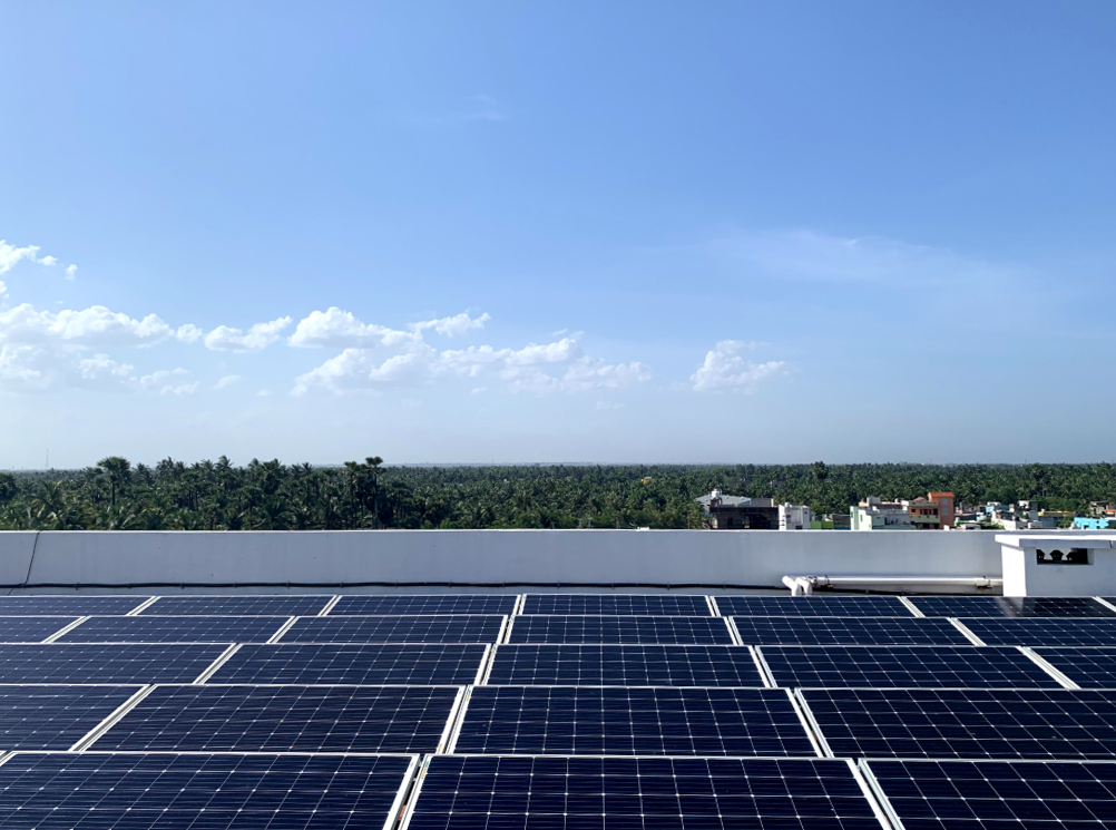 Blue solar panels on a roof. Past the roof's edge, which is smooth and white, there is a dense forest canopy until the horizon line. Above the horizon, the sky is light blue, with a few puffy clouds on the left. 