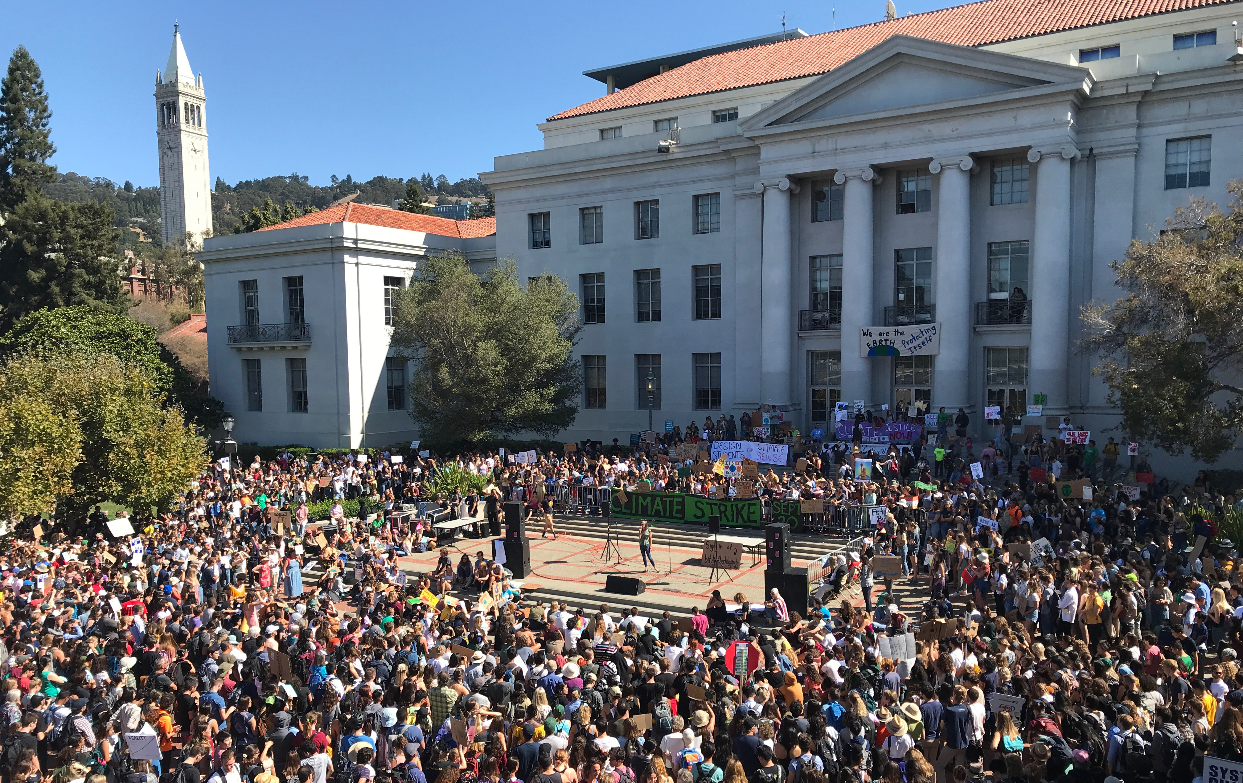 A crowd in front of Sproul Plaza for the Climate Strike. A woman is on a stage and the Campanile is in the background.