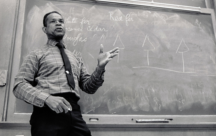 A black and white photo of a man standing in front of a chalkboard in a classroom environment.