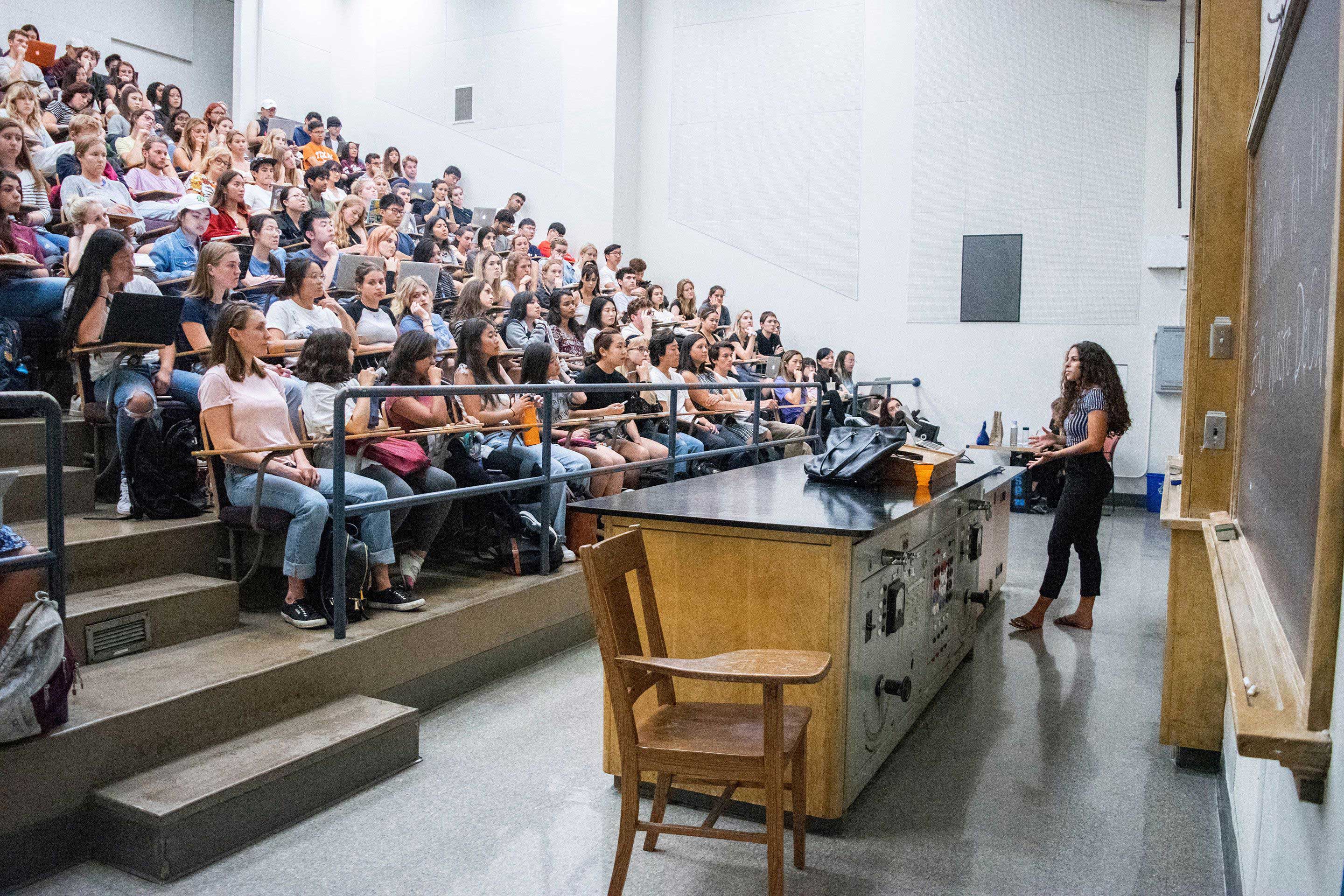 A large lecture hall filled with students and a young woman talking at the front of the room