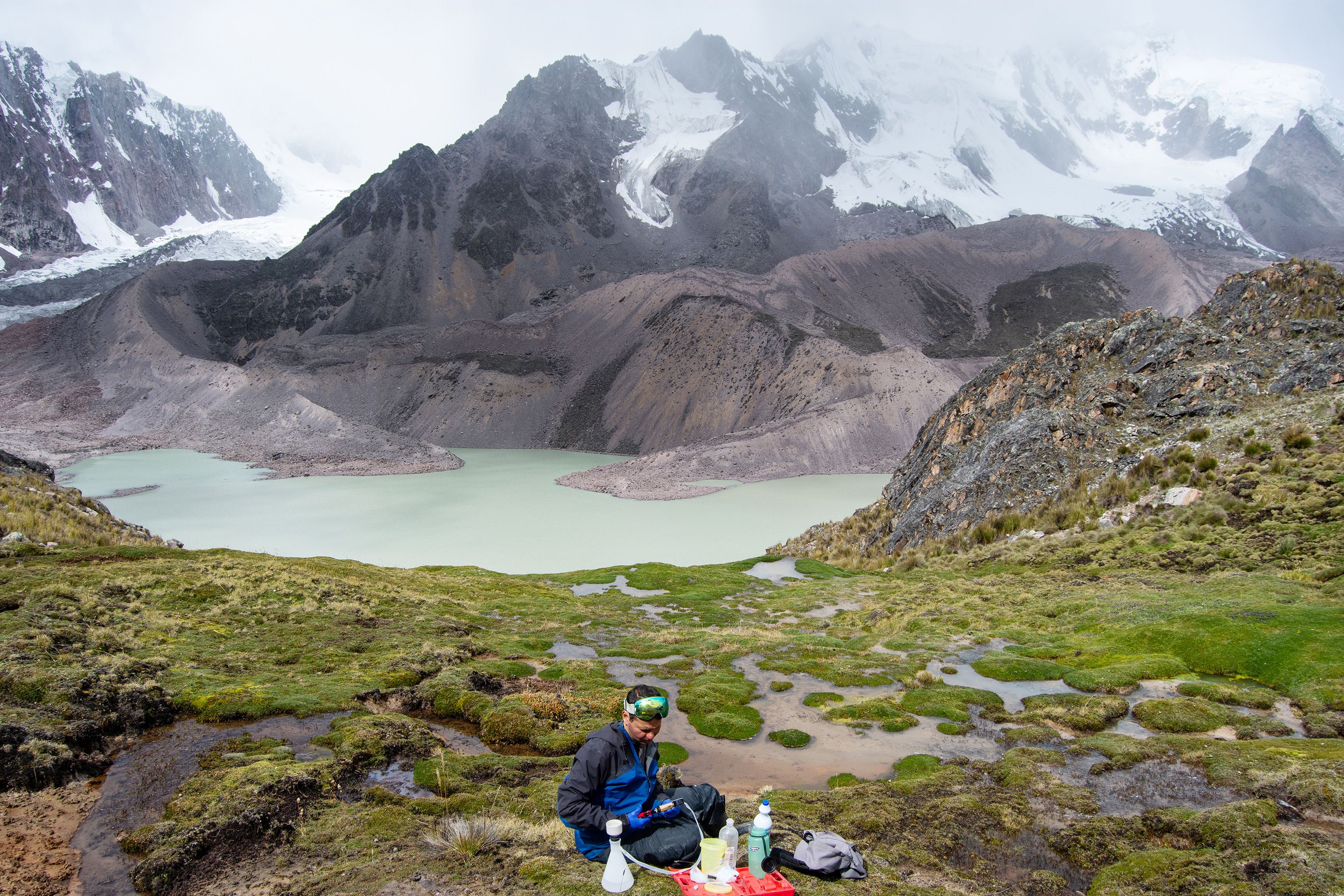 Wide landscape shot, a high altitude mountainous environment takes up most of the image. In the bottom center is a researcher taking measurements on the landscape.