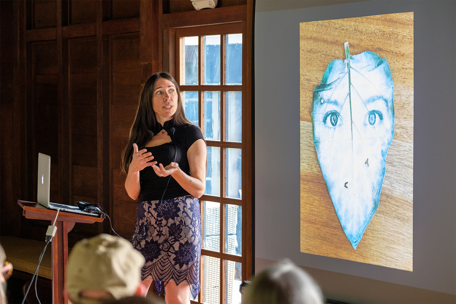 A presenter speaks on a stage, leaf artwork is in the background.