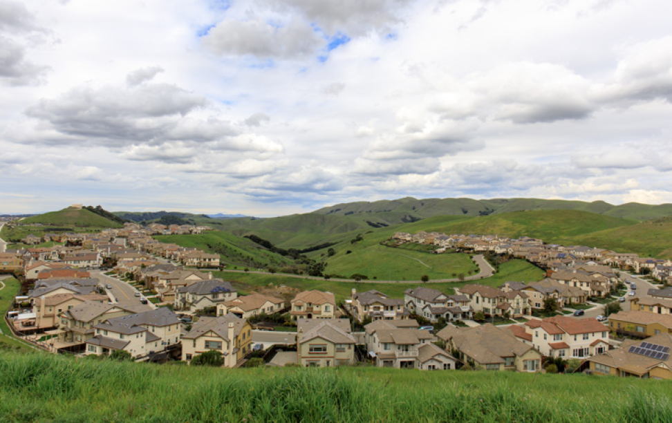 A suburban neighborhood with numerous houses is set against a backdrop of green hills and a cloudy sky.