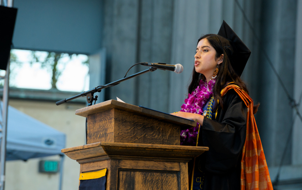 A photo of a woman in graduation regalia speaking while standing at a podium