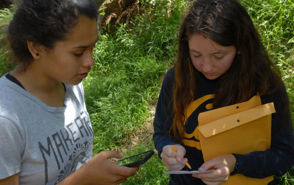 A photo of two young women in a natural environment writing on paper and using a smartphone.