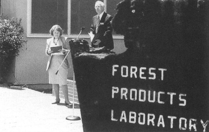 A black and white photo of a man standing at a lectern outdoors. A sign that reads Forest Products Lab is visible in the foreground.