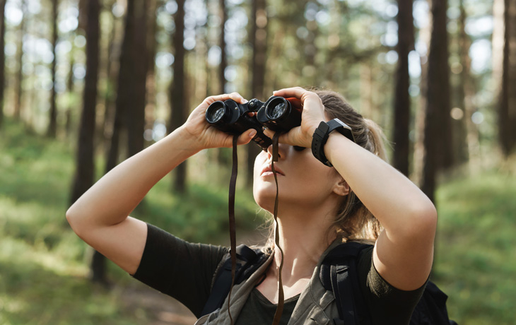 A woman looks through binoculars in a wooded environment.