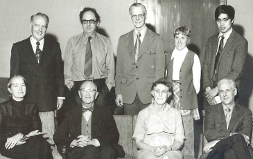 A black and white photo of professors sitting and standing, smiling at the camera