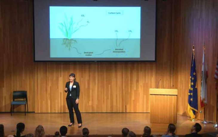 A woman stands on a stage in a suit, presenting to an audience in front of a PowerPoint slide