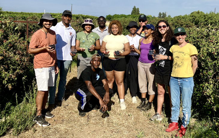 A group of people stand in an agricultural field on a sunny day. They are all holding cups of berries.