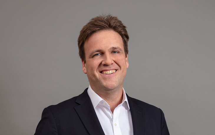 A headshot of a man standing against a grey background. The man is wearing a dark blazer and white shirt, and is smiling at the camera.