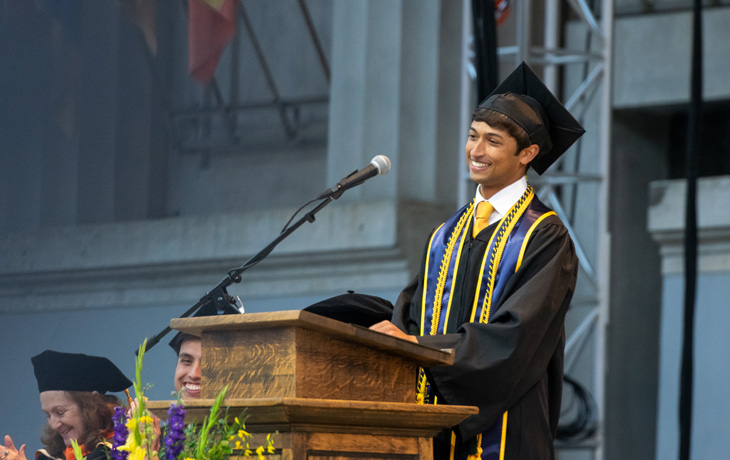 A photo of a man wearing graduation regalia smiling from behind a lectern.