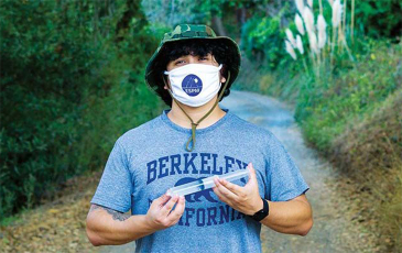 Robin D. Lόpez in front of a forest settting with a fabric face mask covering face, holding scientific equipment.