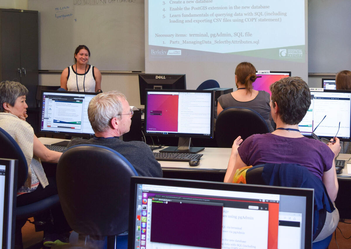 A workshop with participants sitting at computers looking at an instructor with a projection behind her